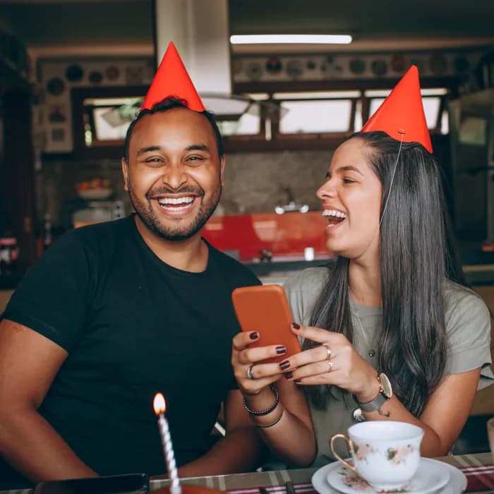 A couple in party hats laughing at something on a phone