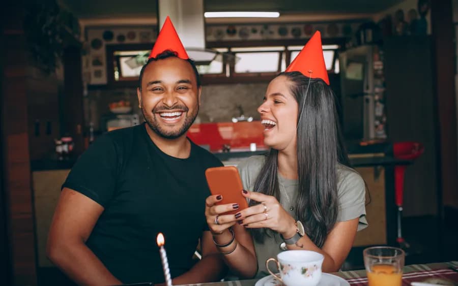 A couple in party hats laughing at something on a phone